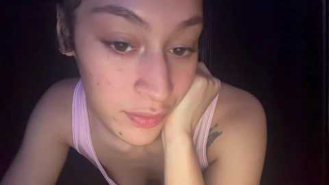 Media: Video of a young Asian woman with light skin, wearing a pink striped tank top, resting her chin on her hand, displaying a tattoo on her upper arm. Dark background.