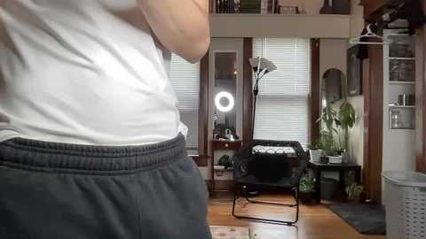 Media: A video of a person wearing a white tank top and gray sweatpants, standing in a cozy, well-lit living room with a black folding chair, potted plants, and a ring light on a desk.