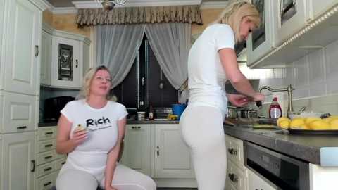 Media: Video of two women in a modern kitchen. One, blonde in a white T-shirt, sits eating a cookie. The other, also blonde, is washing dishes in white leggings.