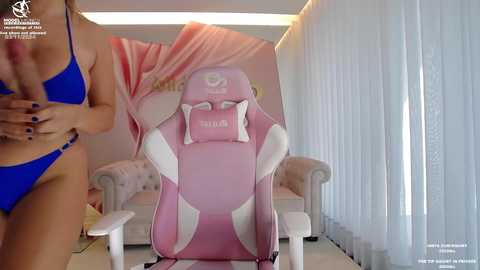 Media: A video shows a woman in a blue bikini, standing next to a pink gaming chair in a brightly lit room with white vertical blinds.