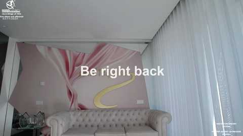 Media: Video of a modern living room with a pink floral wall mural, beige tufted sofa, sheer white curtains, and a \"Be right back\" text overlay.