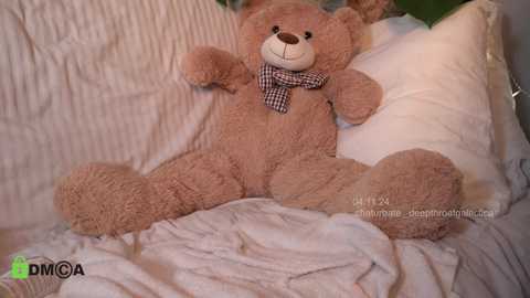 Media: Video of a large, brown teddy bear with a checkered bow tie lying on a soft, white, textured blanket, in a cozy, softly lit bedroom setting.