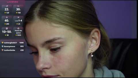 Media: A close-up video of a young woman with fair skin and light brown hair, wearing a grey sweater, sitting in a dimly lit room. Social media notifications overlay the image.