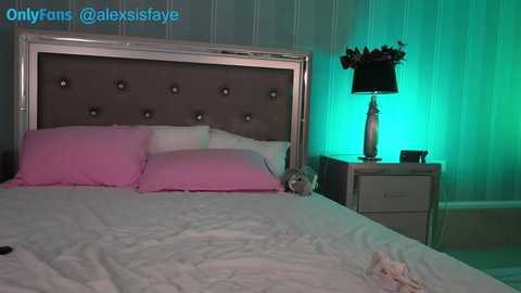 Media: Video of a modern bedroom with a tufted gray headboard, pink pillows, white sheets, teal wall, and a nightstand with a lamp and a stuffed animal.