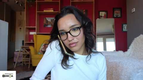 Media: Video of a young woman with long dark hair, wearing black-rimmed glasses and a white top, seated in a modern living room with red walls, wooden shelving, a yellow armchair, and a white furry rug.