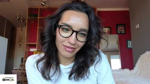 Media: Video of a woman with wavy dark hair, wearing black-framed glasses, a white shirt, and a subtle smile, standing in a cozy, red-walled living room with wooden shelving and framed artwork in the background.