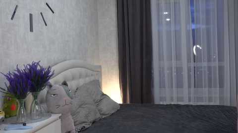 Media: Video of a cozy bedroom with a plush white headboard, gray bedding, a white nightstand with a glass vase of purple flowers, and sheer white curtains.