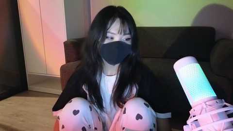 Media: Video of an Asian woman with long black hair, wearing a black mask, white pajamas with heart patterns, and a black top, kneeling on a beige carpet in a dimly lit room, holding a white and blue light-up device.