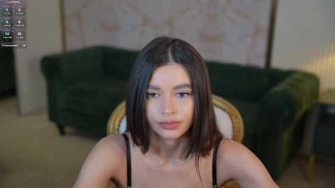 Media: Video of a young woman with straight black hair, fair skin, and light makeup, wearing a black spaghetti-strap top, sitting on a green velvet couch in a softly lit living room.