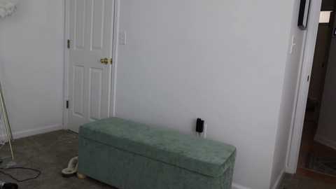 Media: Video of a bare, white-walled room with a closed white door, a mint-green tufted ottoman, and scattered white shoes on the gray carpet.