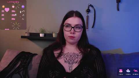 Media: A video of a fair-skinned woman with long dark hair, wearing glasses and a black lace top, with intricate tattoos on her chest, sitting on a couch in a dimly lit room.