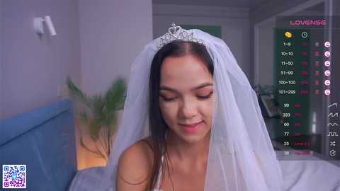 Media: Video of a fair-skinned woman with dark hair, wearing a white veil and a sparkling tiara, smiling in a dimly lit room with a blue couch, a green plant, and a digital display.