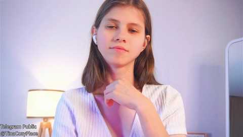 Media: Video of a young Caucasian girl with straight brown hair, wearing a white V-neck shirt, sitting indoors near a lamp, looking contemplative.