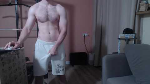 Media: Video of a shirtless, muscular man in white shorts standing in a dimly lit room with peach walls and a grey couch.