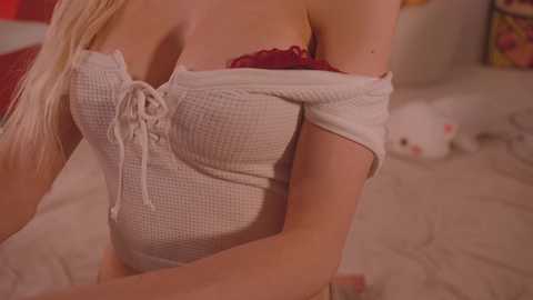 Media: Video of a light-skinned woman with long blonde hair, wearing a white, off-shoulder, ribbed knit top with a red lace bra visible, sitting on a bed with a white, patterned comforter. A white bunny toy is in the background.