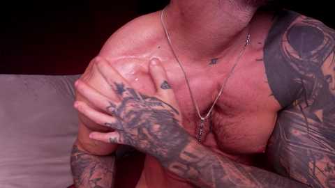 Media: Video of a muscular, tattooed man with light skin, wearing a necklace, gripping his right shoulder, in a dimly lit room with a gray couch.