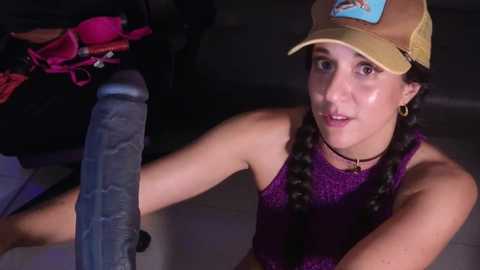 Media: Video of a young woman with fair skin and long brown hair in braids, wearing a yellow cap and purple tank top, holding a large, realistic dildo.