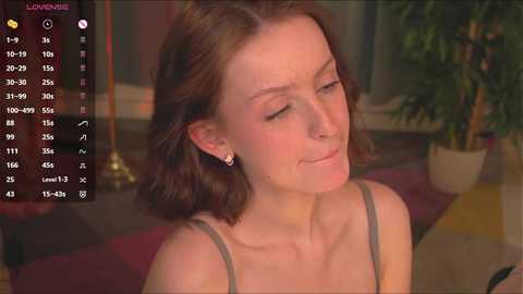 Media: Video of a woman with shoulder-length brown hair, wearing a gray spaghetti-strap top, sitting in a cozy indoor setting with a potted plant and dim lighting.