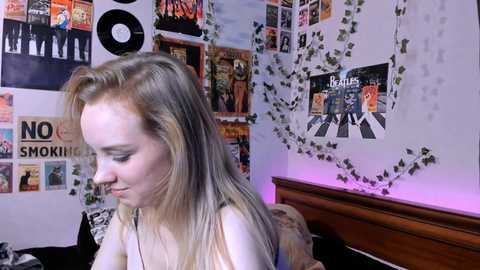 Media: A video of a blonde young woman with pale skin and a slim build, wearing a white top, in a colorful, cluttered bedroom with posters and string lights.