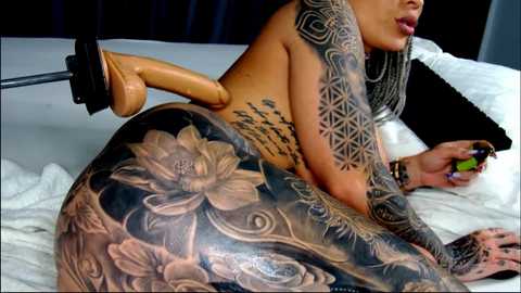 Media: Video of a nude, heavily tattooed woman with intricate floral and geometric designs on her back and arm, lying on a white bed with a black pillow, wearing a tan dildo harness.