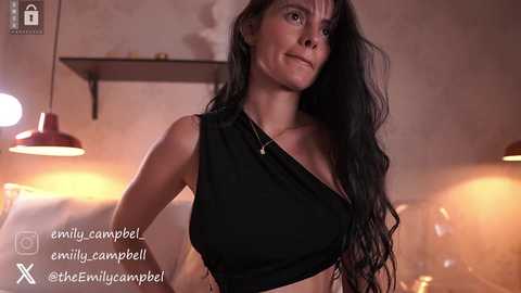 Media: Video of a slender, dark-haired woman with fair skin, wearing a black off-shoulder top, standing in a dimly lit room with warm, ambient lighting.