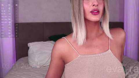 Media: Video of a light-skinned, blonde-haired woman with a slim physique and medium-sized breasts, wearing a beige, ribbed, sleeveless top, in a bedroom with a gray upholstered headboard, white pillows, and sheer purple curtains.