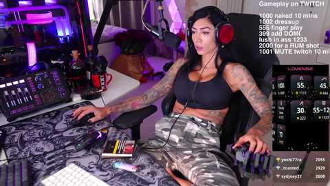 Media: Video of a tattooed, curvy woman with long black hair, wearing a black sports bra and camo pants, playing a video game on a dark gaming setup.