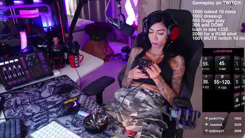 Media: A video of a tattooed woman in camo pants, wearing headphones, playing a video game. She sits at a desk cluttered with gaming equipment and drinks.