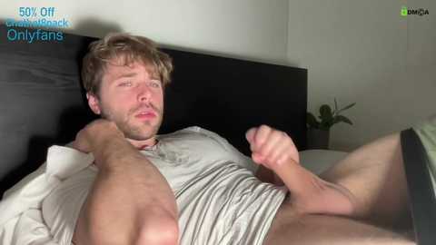 Media: Video of a young, muscular, fair-skinned man with light brown hair, wearing a grey shirt, lying on a bed, with a large erect penis visible.