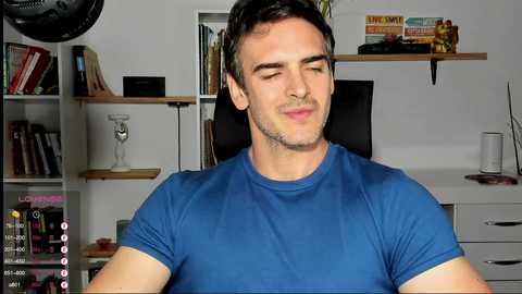 Media: Video of a young man with short dark hair, wearing a blue t-shirt, smiling, in a modern, organized living room with shelves, books, and a lamp.