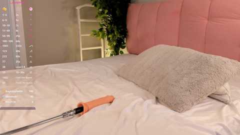 Media: A video of a modern bedroom with a pink upholstered headboard, white bedding, and a textured gray pillow. An orange screwdriver is lying on the bed, with a white ladder and green plants in the background.