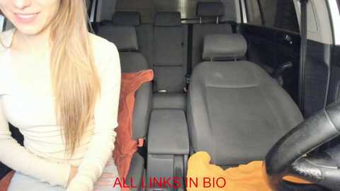 Media: Video of a woman with long blonde hair, smiling, sitting in the passenger seat of a car with a beige jacket. The car seats are gray and orange blankets are visible in the back.