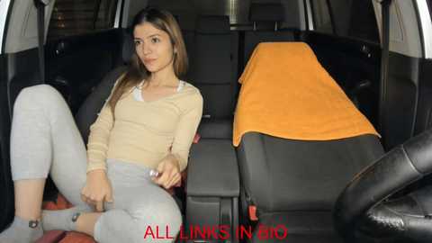 Media: Video of a young woman with long brown hair, light skin, wearing a beige sweater and gray pants, seated in a car with a black seat and orange blanket, looking relaxed.