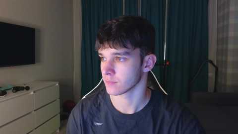 Media: Video of a young man with short dark hair wearing headphones, standing in a dimly lit bedroom with a TV, dresser, and dark curtains.