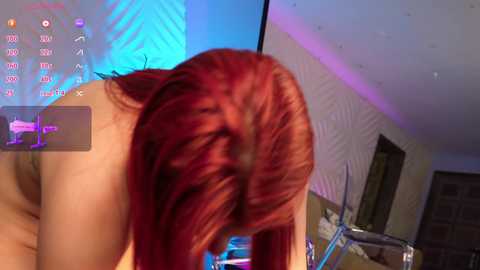 Media: Video of a naked woman with long red hair, bending forward, in a dimly lit room with purple and blue lighting.