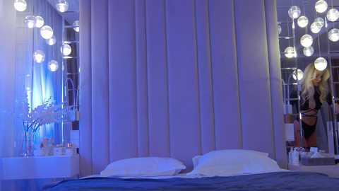 Media: Video of a modern bedroom with a tall, vertical striped headboard, white pillows, and a blue blanket. Hanging spherical pendant lights cast a soft, blue glow. A mirror reflects a blonde woman in a black outfit, holding a phone.