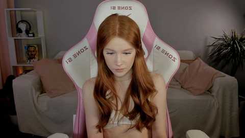 Media: Video of a pale-skinned, red-haired young woman sitting on a pink gaming chair, wearing a white bra, in a cozy living room with a beige couch and shelves.