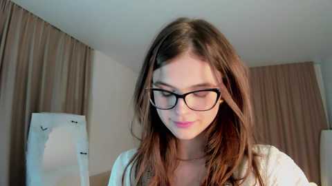 Media: Video of a young Caucasian woman with long brown hair, wearing black-framed glasses, light makeup, and a white shirt, in a beige room with curtains and a white chair.