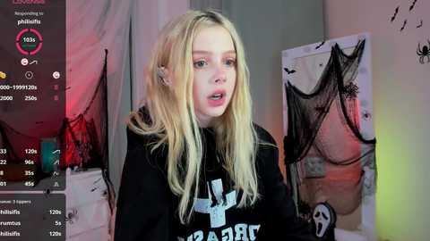 Media: Video of a young, fair-skinned, blonde woman with straight hair, wearing a black sweatshirt, in a dimly lit room with art on the walls, surrounded by a live stream interface.