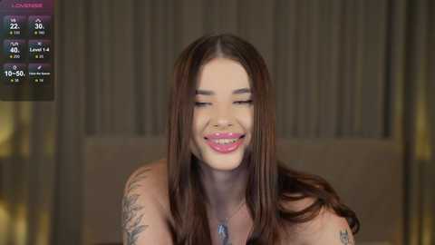 Media: A video of a smiling woman with long, straight brown hair, wearing a necklace, sitting in a dimly lit room with a TV screen showing a live stream.