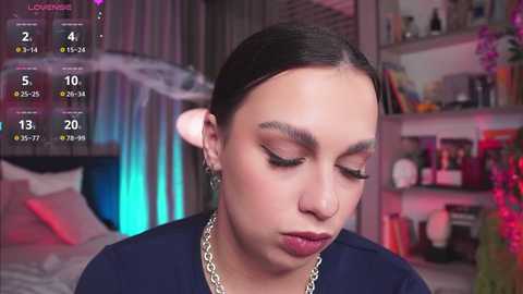 Media: Video of a young woman with medium-brown skin, long dark hair, and thick eyebrows, wearing a black top and silver chain necklace, in a bedroom with a bed, bookshelf, and colorful decor.