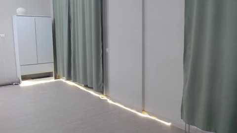 Media: Video of a minimalist, brightly-lit room with a white floor and walls. Light green curtains cover the windows, casting soft shadows on the floor. A white cabinet stands against the left wall.