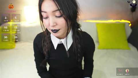 Media: Video of a young woman with pale skin and long black hair in braids, wearing a black sweater over a white collared shirt, and dark lipstick, in a modern room with yellow and white decor.