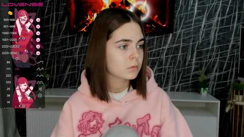 Media: Video of a young woman with straight brown hair, wearing a pink hoodie with cartoon characters, in a room with a fireplace and grey wall.