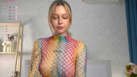 Media: Video of a blonde woman with fair skin, wearing a colorful, sheer mesh top, standing indoors with a white wall and blue curtains in the background.