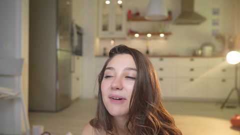 Media: Video of a young woman with long brown hair, eyes closed, smiling, in a modern, well-lit kitchen with white cabinets, stainless steel appliances, and a round pendant light.