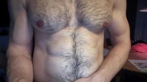 Media: A video of a muscular, bare-chested man with a full beard and hairy chest, holding his belly. The background features a cluttered room with a wooden table and a pink object.