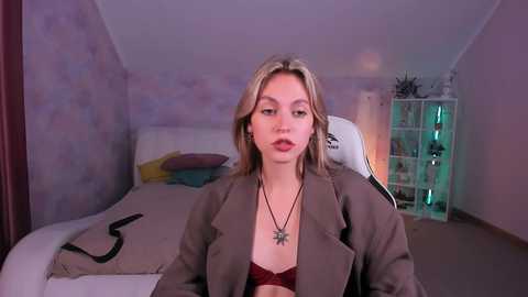 Media: Video of a blonde woman with light skin, wearing a maroon bra and beige blazer, sitting on a white chair in a dimly-lit, pastel-colored bedroom.