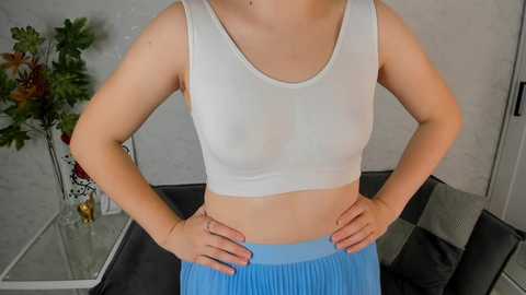 Media: Video of a light-skinned woman with fair skin, wearing a white sports bra and blue pleated skirt, standing with hands on hips in a modern, minimalist room with grey walls and a black table.