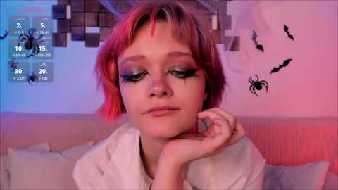 Media: Video of a young woman with short, vibrant red hair and striking makeup, including dark eyeliner, wearing a white shirt. Background features a Minecraft-themed wall with pixel art and animated bats, set in a cozy room with soft lighting.